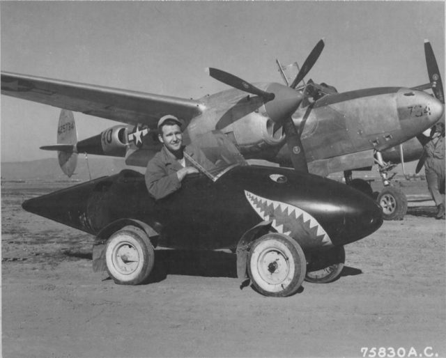 United States Army Air Forces Lockheed P-38L Lightning aircraft ( Serial Number – 44-25734 ) and a ground crew member of the 94th Fighter Squadron 1st Fighter Group, poses in his self-styled auto made from salvaged Lockheed P-38 Lightning parts including a fuel tank with wheels added and a plexiglass windshield. This P-38 while assigned to 1st FG, 71st FS was shot down by AAA near Munich April 15, 1945.