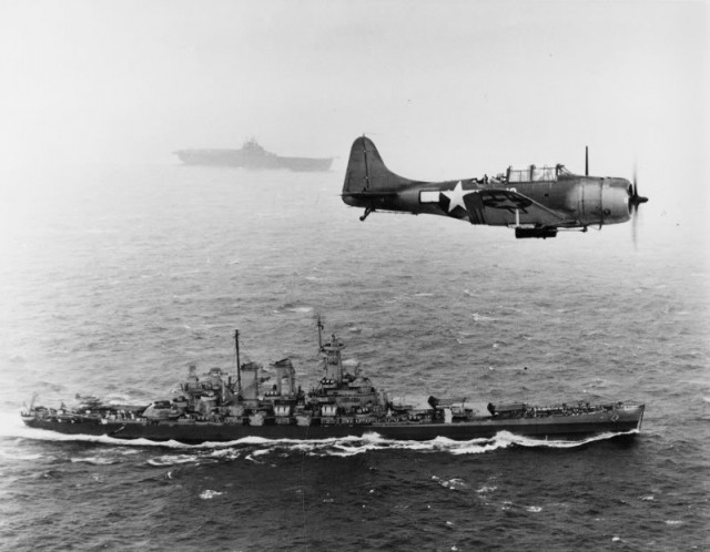 A U.S. Navy Douglas SBD-5 Dauntless of bombing squadron VB-16 flies an antisubmarine patrol low over the battleship USS Washington (BB-56) en route to the invasion of the Gilbert Islands, 12 November 1943. The ship in the background is USS Lexington (CV-16), the aircraft’s home carrier.